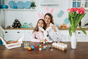Mom and daughter wearing bunny ears are having fun with Easter bunny while sitting on kitchen....