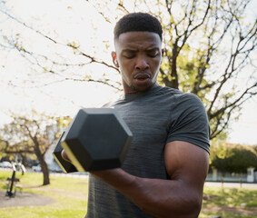 Fit young black man in t-shirt and wireless earphones doing bicep curl with dumbbell in park - 762810100
