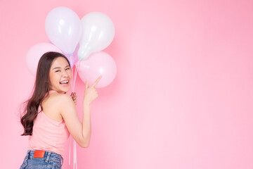 Excited cheerful asian woman holding balloons and pointing finger smiling with toothy standing over isolated pink background. Joyful teenager girl with pastel balloons shocked  amazed expression.