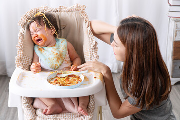 Unhappy mixed race toddler child messy tomato sauce on face crying with mother sitting together...