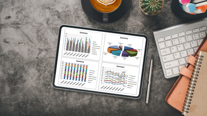 Tablet on a gray desk showing business summary graphs, next to a coffee, keyboard, and stationery.