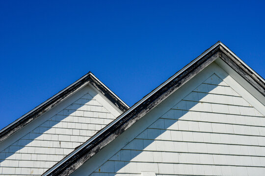 Multiple peaked and layered roofs on a white vintage wooden house. The eave is painted black. The old building has narrow wood cedar clapboard siding painted white. The background is a bright blue sky