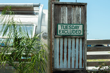 Tuesday excluded sign with green lettering and a cream colored background. The small wooden sign is attached to a large board blind next to a silver RV camper. There's a rolled canvas awning attached.