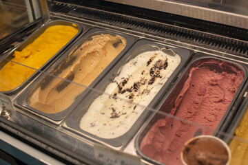 Multiple metal trays containing dairy mango, cherry, raspberry, mocha, vanilla, chocolate and caramel creamy gelato ice cream for sale in an ice-cream parlor. The various flavors are for round scoops.