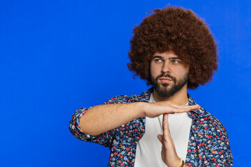 Tired serious upset man with Afro hairstyle wig showing time out gesture, limit or stop sign, no pressure, I need more time, take a break, relax rest, help. Guy isolated on blue background. Copy-space