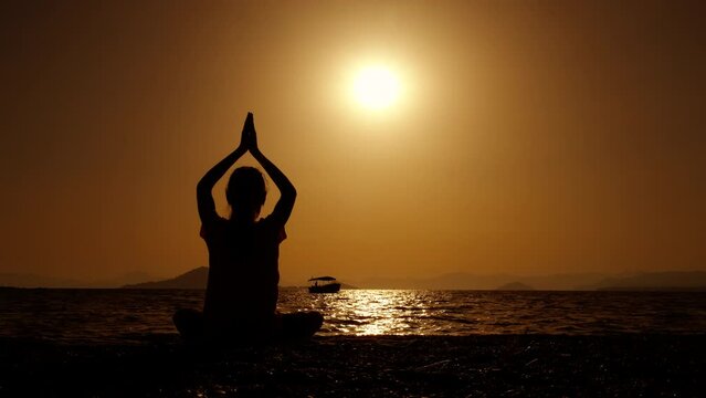 Girl silhouette in prayer gesture. A view of cute teen silhouette in namaste pose relaxing on the beach during nightfall time. A concept of sporty lifestyle during summer vacation.