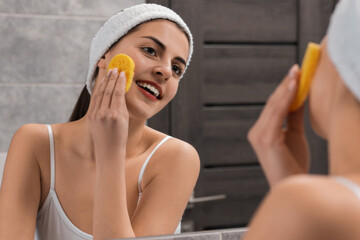 Young woman with headband washing her face using sponge near mirror in bathroom