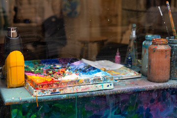 A close-up look at an artist's workspace with a messy paint palette and stained jars filled with...
