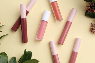 Different lip glosses, green leaves and flowers on pale yellow background, flat lay