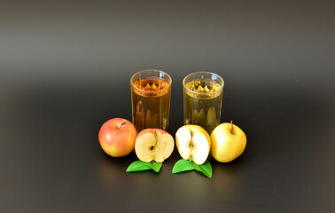 Two glasses with different fruit juices on a black background, next to pieces of ripe red and yellow apples with leaves.