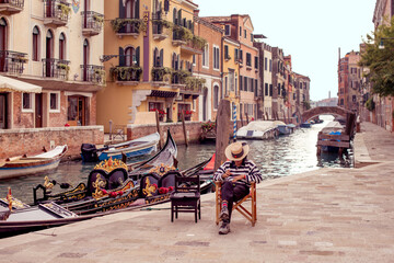 A gondolier in Venice is waiting for customers on the background of a gondola. The gondolier is bored against the background of the view of the Venetian canals - obrazy, fototapety, plakaty