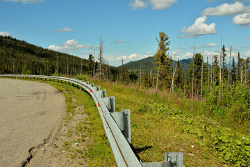 A fragment of an asphalt road with a fence on top of a hill overlooking the picturesque mountain ranges on a sunny summer day.