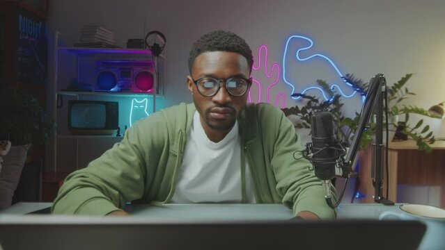 Young African American man sitting at desk in modern video studio with neon light and surfing the Internet on laptop when working on content plan for social media blog. Zoom shot