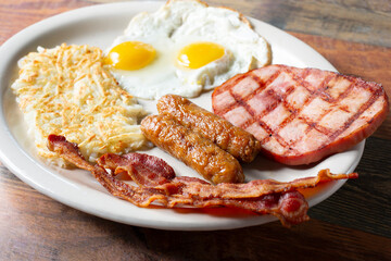 A view of a breakfast plate, featuring eggs, hash browns, ham, sausage and bacon.