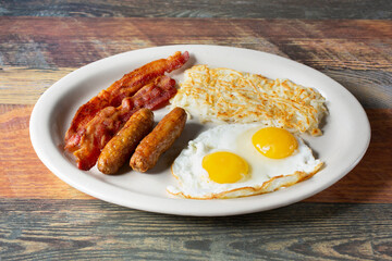 A view of a breakfast plate, featuring fried eggs, hash browns, sausage and bacon.
