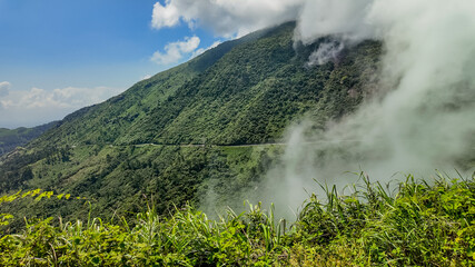 Lush green mountain slopes with low-hanging clouds, winding road in the distance, suitable for environmental or travel-themed backgrounds with space for text