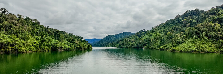 Fototapeta na wymiar Panoramic view of a tranquil tropical lake with lush green rainforest under an overcast sky, ideal for environmental themes and nature backgrounds with copy space, Earth day concept