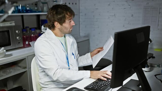 A caucasian man with a beard reads a document while working on a computer in a laboratory setting.