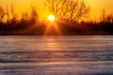 Spring landscape. Sunrise of the solar disk over the northern icy river, on the bare winter floodplain forests. Northeast of Europe - obrazy, fototapety, plakaty
