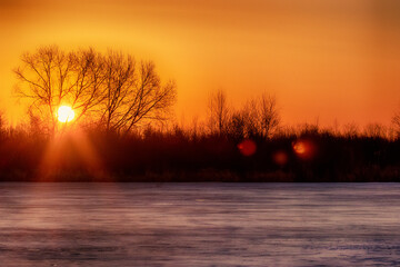 Spring landscape. Sunrise of the solar disk over the northern icy river, on the bare winter floodplain forests. Northeast of Europe - obrazy, fototapety, plakaty