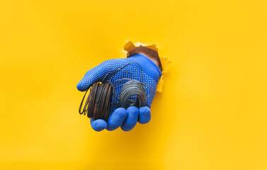 A man's hand in a blue knitted glove holds a two coil of steel wire. Torn hole in yellow paper. The concept of a worker, a labor migrant, a master of his craft. Copy space. - Powered by Adobe