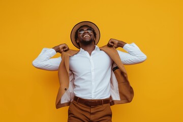 Joyful African man in stylish brown hat and sunglasses pulling off jacket on a vibrant yellow background, embodying confidence and happiness - obrazy, fototapety, plakaty