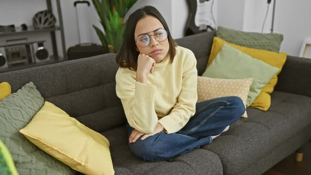 Bored and tired, a young hispanic woman wearing glasses, depicts a sad expression of depression at home. arm crossed, thinking deeply about her problems in her living room.