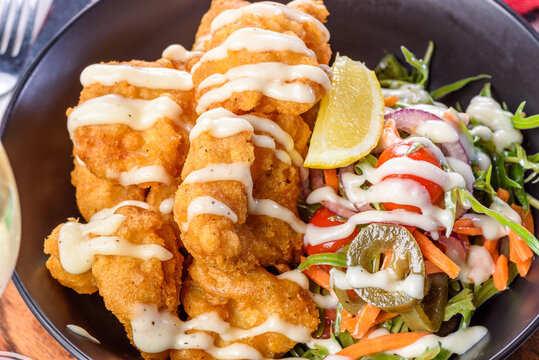 A serving of deep-fried fish pieces with a white sauce and salad to the side.
