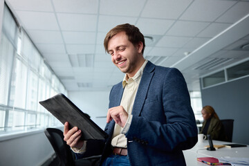 Happy young businessman in businesswear using digital tablet by desk with coworkers in office - 762954951