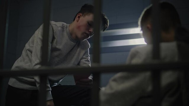Caucasian teenage prisoner plays cards in prison cell with other inmates. Young criminals serve imprisonment terms in jail. Juvenile detention center or correctional facility. View through metal bars.