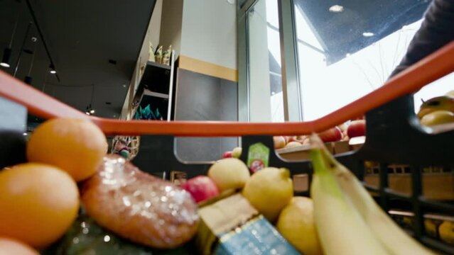 Shopaholic going through rows of grocery shop and adding variety of products inside black and orange cart. Close up view on content of shopping basket getting completely filled with edible items.