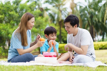 Happy cheerful Asian family with father, mother, and little son enjoy picnic together in a weekend at a park.