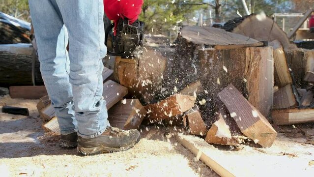 Slow motion while using chainsaw to cut slabs of oak into smaller pieces sending shavings toward camera while preparing firewood for winter season.