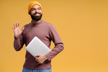 Handsome smiling bearded African American man wearing stylish yellow hat, waving hand holding laptop