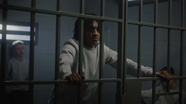 Multiethnic teenagers behind metal bars in prison cell look at camera. Young prisoners, criminals serve imprisonment terms for crimes in jail. Youth detention center or correctional facility. Portrait