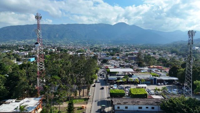 Cars on main road of Caribbean town with transmission tower and park trees. Mountain range and cityscape of Jarabacoa, Dominican Republic in Background. Aerial forward birds exe shot.
