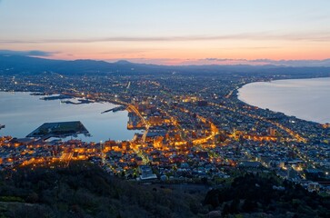 A beautiful panoramic view of Hakodate at dawn, with sparkling city lights in downtown flanked by sea shores on both sides and golden twilight sky on distant horizon before sunrise, in Hokkaido, Japan
