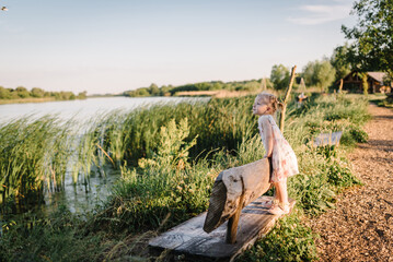 Kid on a bench on the shore of the lake at sunset. Girl in the summer park. Child walking outdoors. A family walk with children.