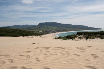 "Dunes of Bolonia Beach with Tarifa in the  Sand dunes roll gently toward a serene beach that hugs the coastline with lush greenery and clear blue skies overhead.