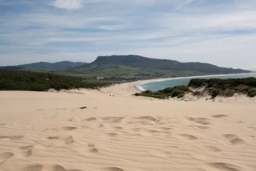 Fotobehang Bolonia strand, Tarifa, Spanje "Dunes of Bolonia Beach with Tarifa in the  Sand dunes roll gently toward a serene beach that hugs the coastline with lush greenery and clear blue skies overhead.