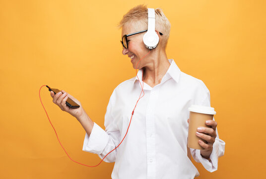Cheerful pensioner woman with short haircut listens to music and holds a glass of coffee, dances on a yellow background