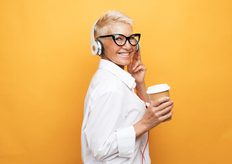 senior woman with short hair listening to music with headphones and holding takeaway coffee