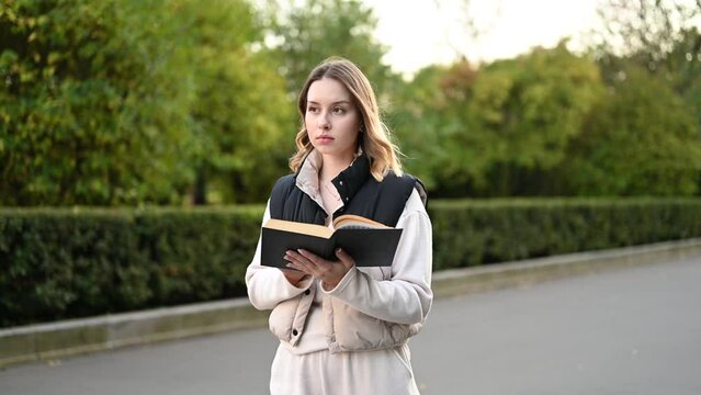 Young woman walking through the park and reading a paper book