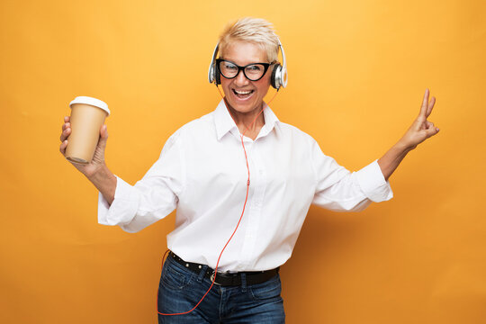 Cheerful pensioner woman with short haircut listens to music and holds a glass of coffee, dances on a yellow background