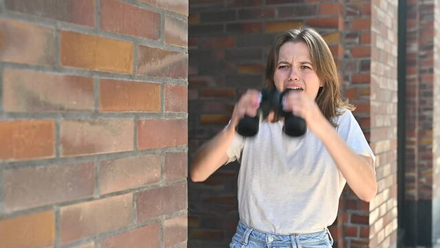 A young woman peeks funny around the corner with binoculars and spies on someone, making funny faces