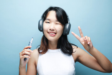 Cheerful Asian teenage girl listens to music on headphones and shows a victory sign.