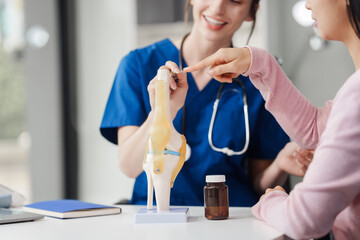 Caucasian female doctor explain to asian female patient using knee bone model at desk in medical...