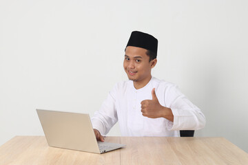 Portrait of excited Asian muslim man in koko shirt with skullcap working on his laptop during fasting on ramadan month, showing thumb up hand gesture. Isolated image on white background