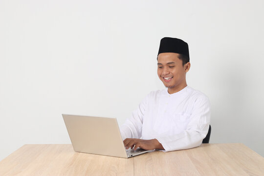 Portrait of excited Asian muslim man in koko shirt with skullcap working on his laptop during fasting on ramadan month. Isolated image on white background