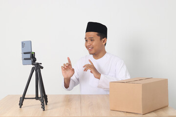Portrait of excited Asian muslim man in koko shirt with skullcap promoting his product on live streaming session. Online shopping with smartphone concept. Isolated image on white background